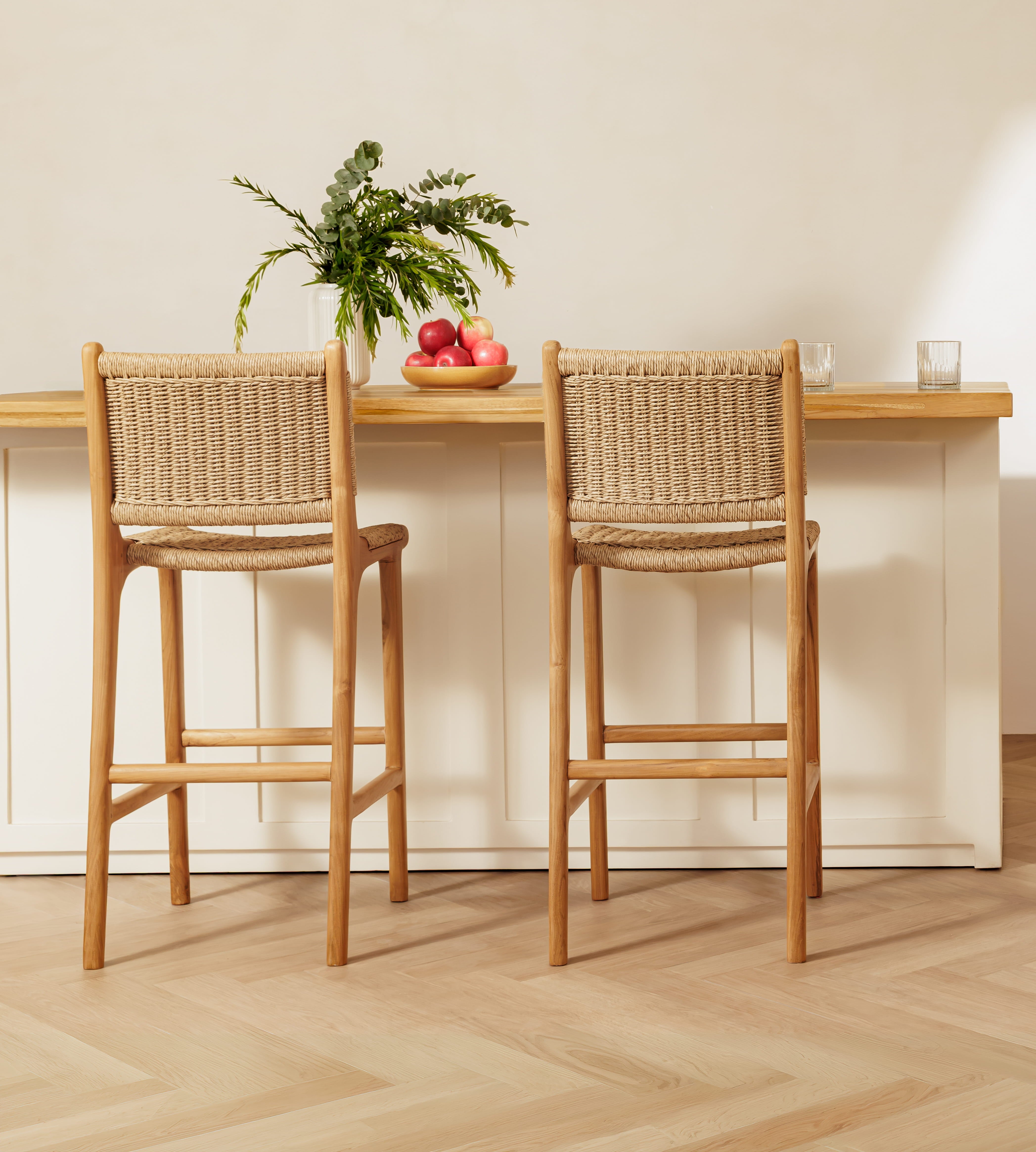 Two wooden bar stools with woven natural-fiber seats and backs, positioned in front of a light wood counter in a bright, modern interior with a pale wood herringbone floor.