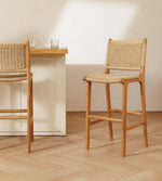 Two rattan bar stools with woven rattan seats and backs, shown in a bright, modern kitchen setting with a light wood table and white cabinets.