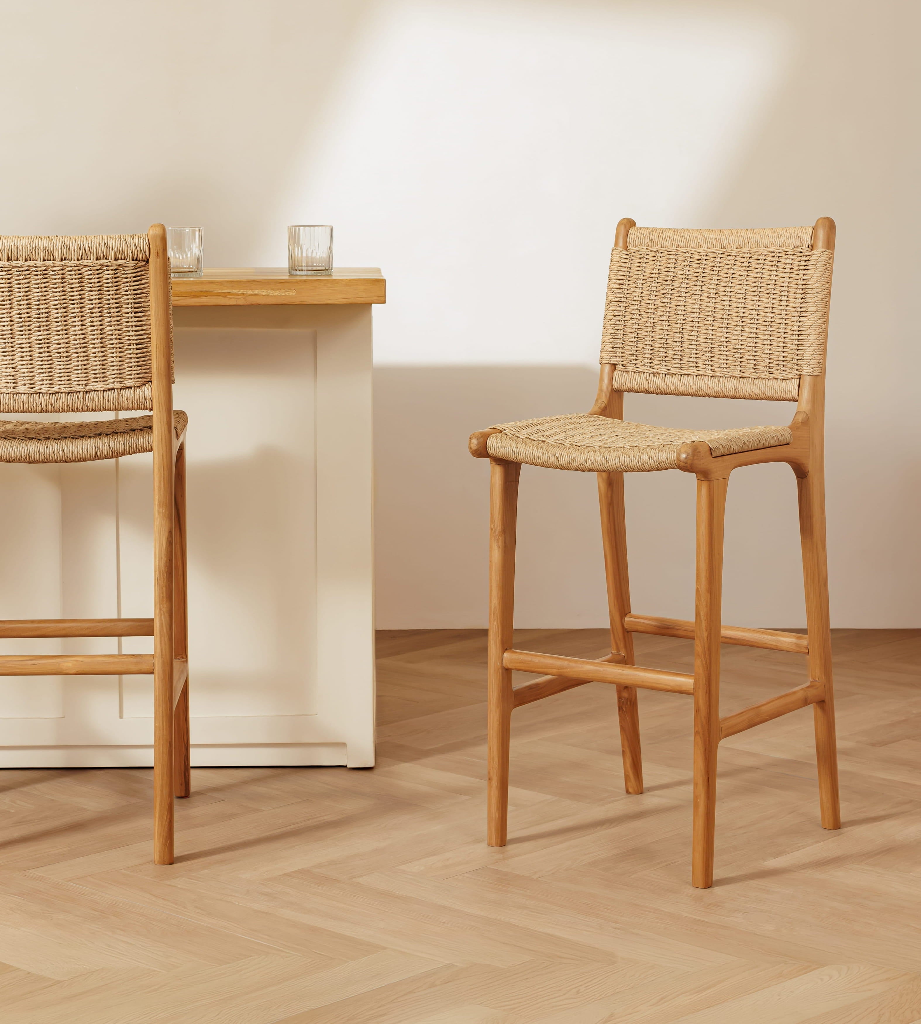 Two rattan bar stools with woven rattan seats and backs, shown in a bright, modern kitchen setting with a light wood table and white cabinets.