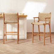 Two wooden bar stools with woven rope seats and backs are shown in a bright, modern kitchen or dining area with light wood flooring and a white counter.