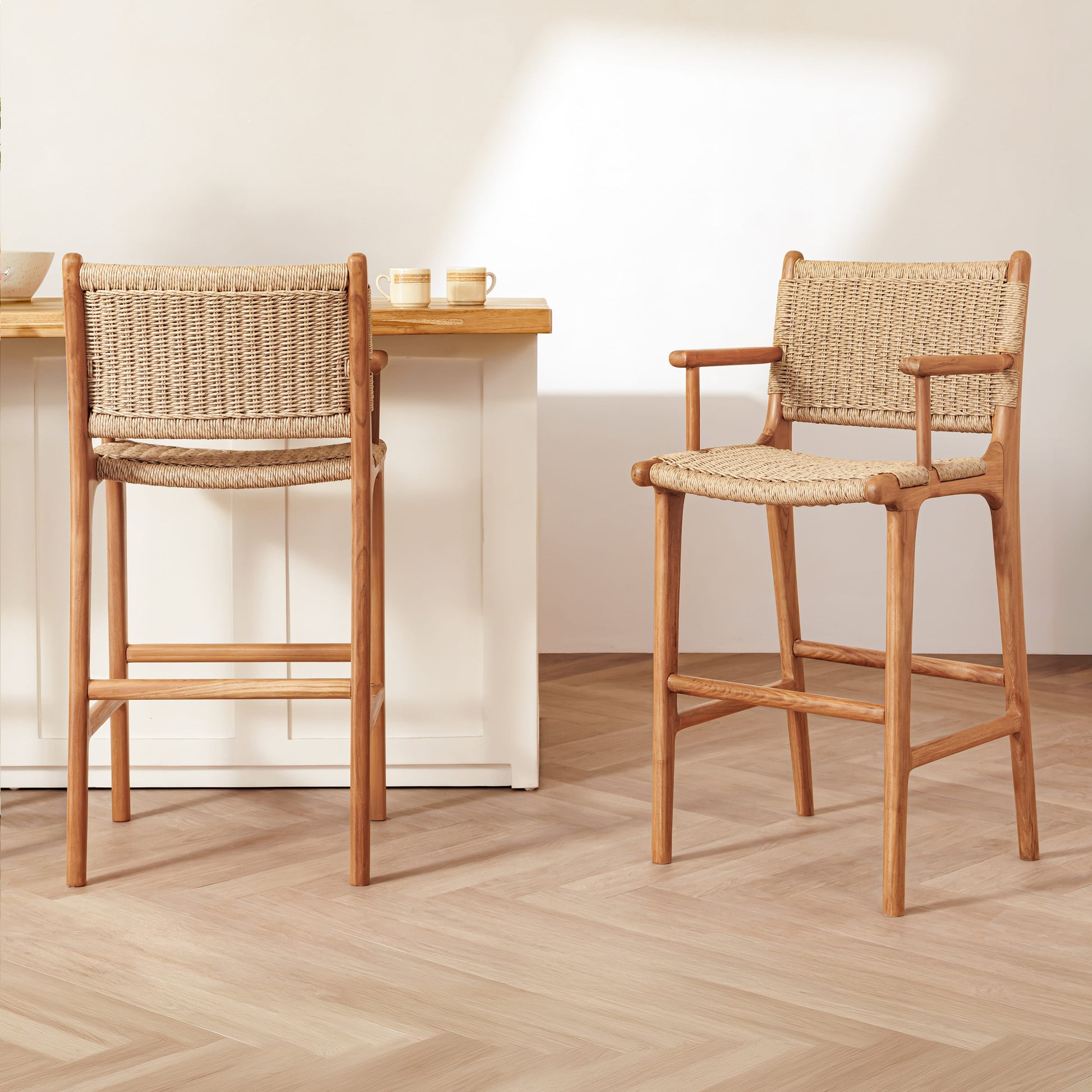 Two wooden bar stools with woven rope seats and backs are shown in a bright, modern kitchen or dining area with light wood flooring and a white counter.