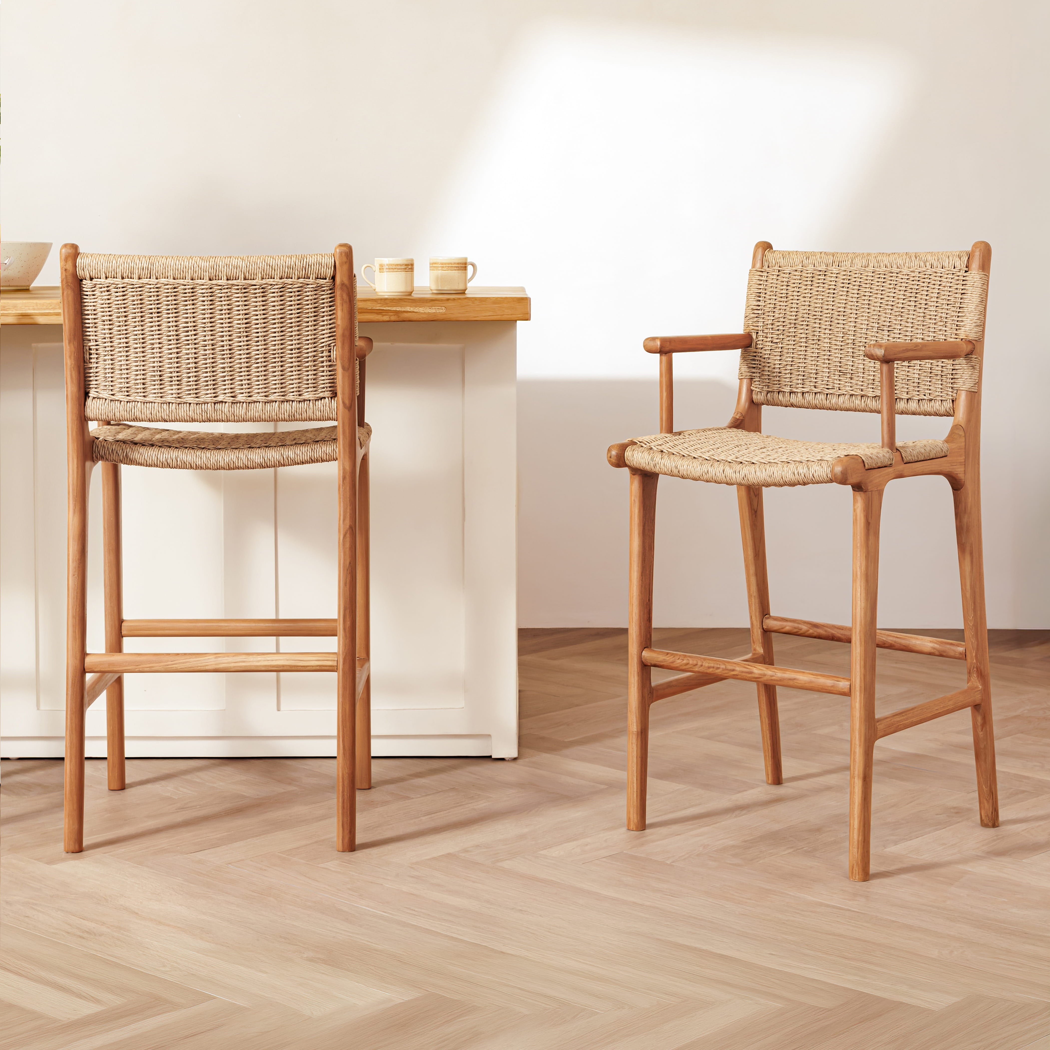 Two wooden bar stools with woven rope seats and backs are shown in a bright, modern kitchen or dining area with light wood flooring and a white counter.