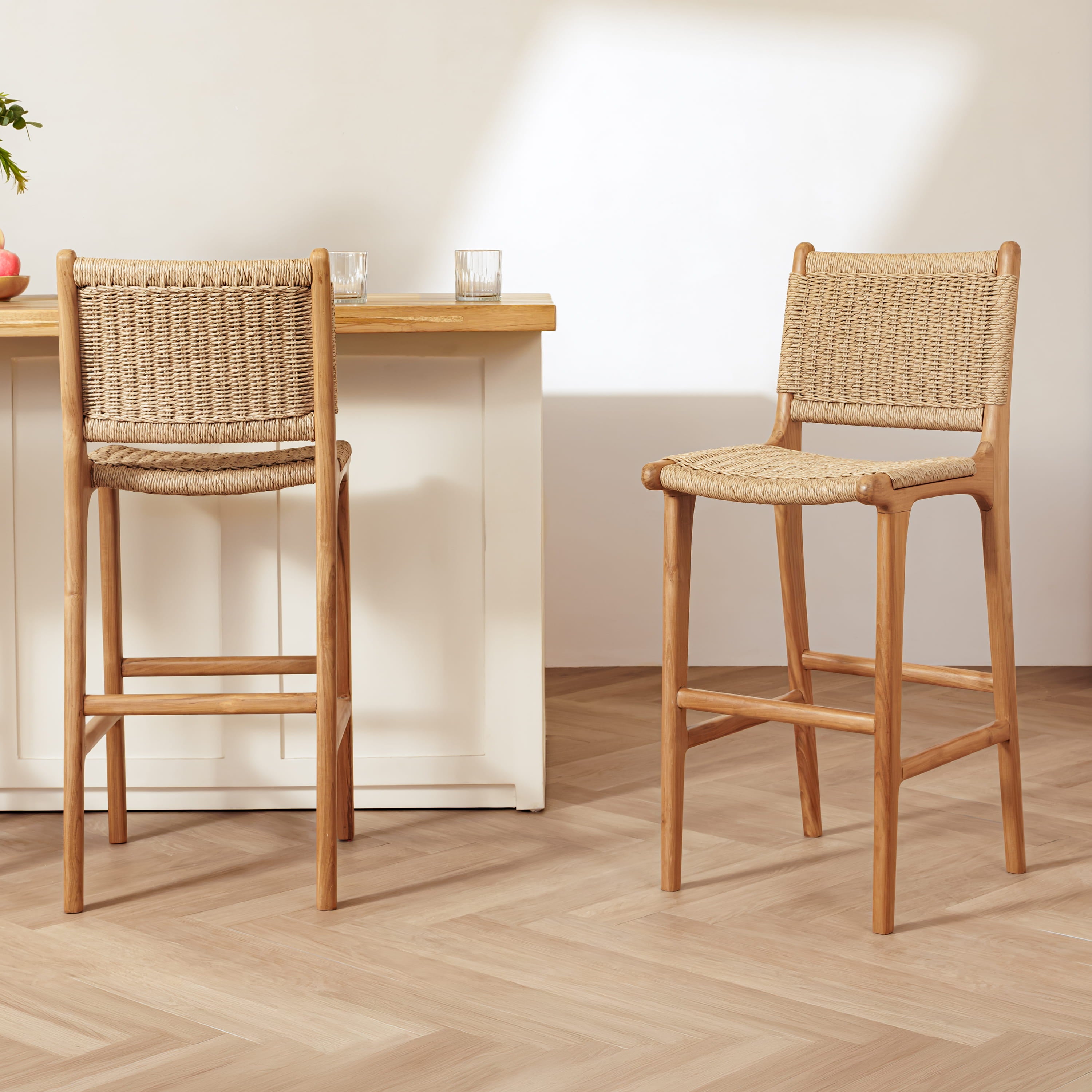 Two rattan bar stools with woven rattan seats and backs, shown in a bright, modern kitchen setting with a light wood countertop and herringbone floor.